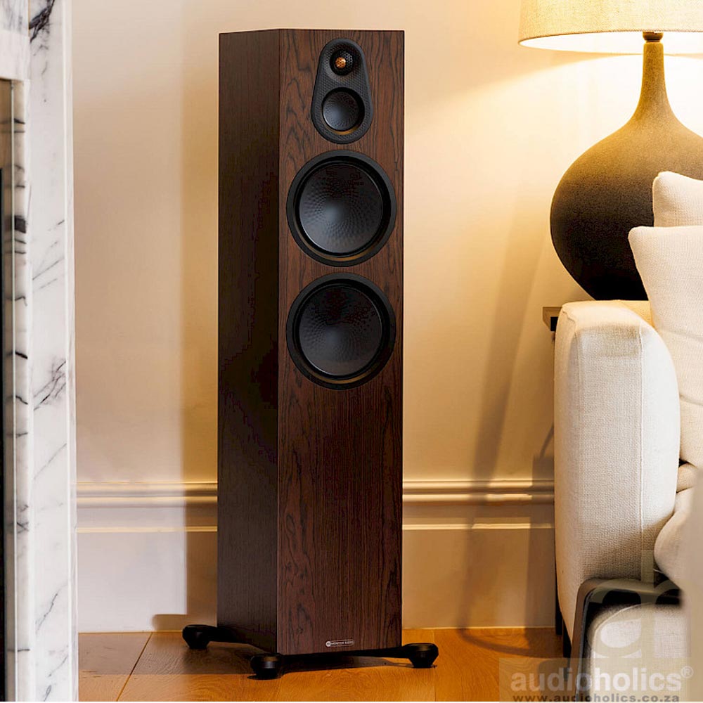 Monitor-Audio-Silver-500-Main - Audioholics South Africa Tall wooden floor-standing speaker with three driver units (two large woofers and a tweeter) in a living room beside a sofa and lamp, on a wheeled base.