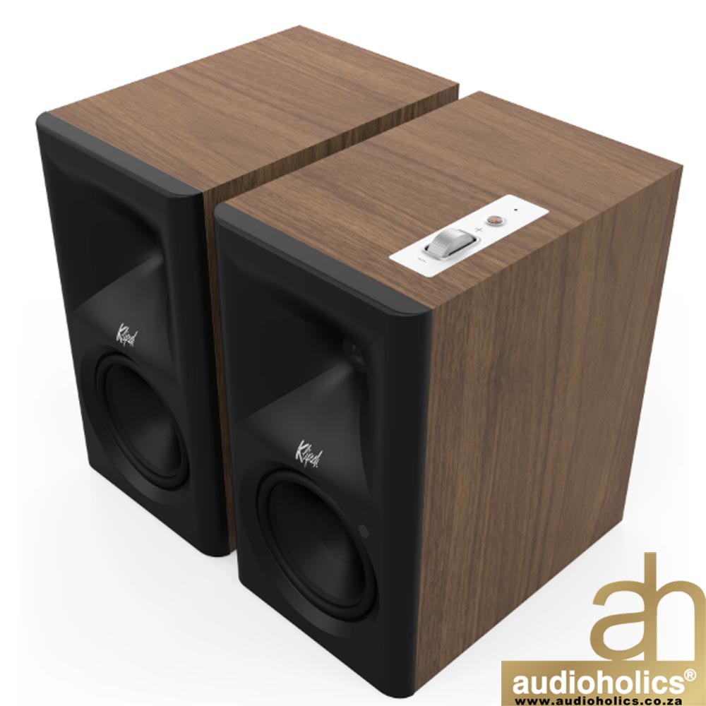 Klipsch-The-Fives-II-Main---Walnut-1 - Audioholics South Africa Pair of walnut-finish bookshelf speakers with black front baffles, shown from an angled top view; one speaker has a white control panel on its top surface and Audioholics logo in the corner.